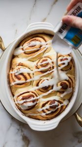 a freshly baked cinnamon roll casserole in a white oval baking dish being drizzled with white icing from a packet