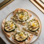 flatbreads being topped with grilled zucchini slices, crumbled feta, and lemon zest on a plate