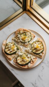 flatbreads being topped with grilled zucchini slices, crumbled feta, and lemon zest on a plate