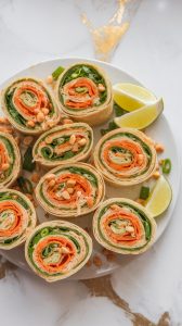 Ultra close-up of Thai Chicken Peanut Pinwheels sliced into bite-sized rounds, showing the layers of shredded chicken, creamy peanut sauce, shredded carrots, and fresh cilantro wrapped in a soft flour tortilla. Some are stacked and some are fanned out on a white plate, sprinkled with chopped peanuts and green onions. Garnished with lime wedges. White marble counter with hints of gold, with all-natural lighting, taken with an iPhone 15 Pro, top-down shot used by recipe bloggers.