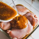 marinade being poured over raw chicken breasts in a shallow glass dish, coating them completely.