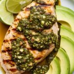 grilled chicken breast plated with chimichurri spooned over the top and fanned avocado slices on the side, garnished with lime wedges.