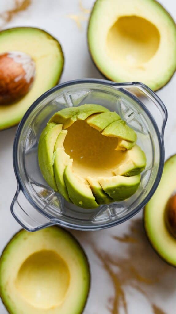 avocado halves with flesh scooped into a blender cup, bright green and soft. White marble counters with hints of gold.
