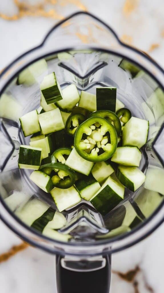chopped cucumber and jalapeño pieces inside a blender cup, fresh and vibrant. White marble counters with hints of gold