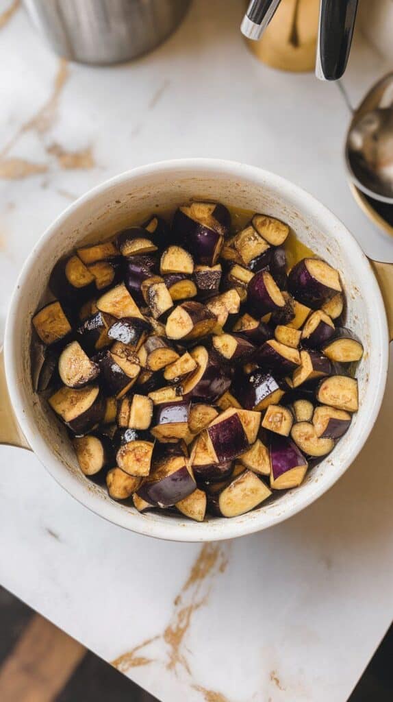 cubed eggplant glistening with olive oil and seasoning, mixed in a large white mixing bowl. White marble counters with hints of gold.