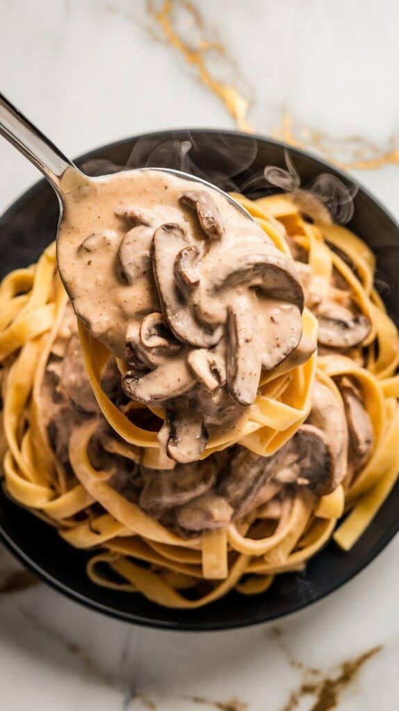 creamy mushroom sauce being spooned over tagliatelle pasta, thick and clinging to the noodles with steam rising.