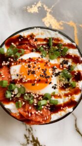 inished kimchi egg cottage cheese bowl being drizzled with soy sauce and sprinkled with green onions, sesame seeds, and gochugaru, vibrant colors and textures showing.