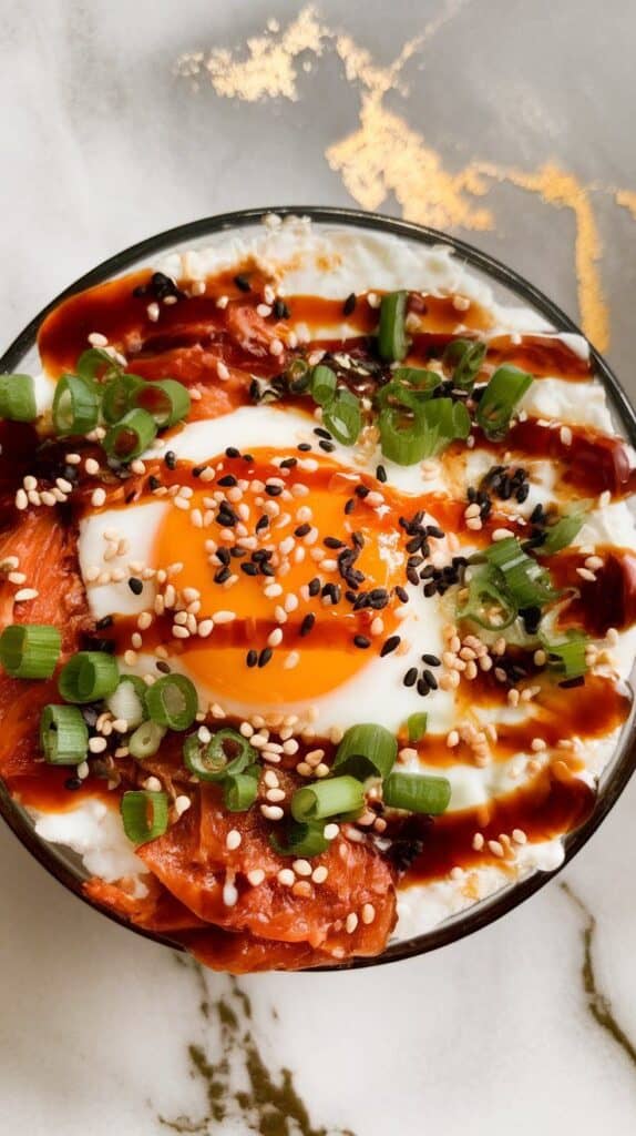 inished kimchi egg cottage cheese bowl being drizzled with soy sauce and sprinkled with green onions, sesame seeds, and gochugaru, vibrant colors and textures showing.