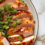 sliced chicken breast on a plate drizzled with thick sriracha honey garlic glaze, topped with chopped green onions and sesame seeds.