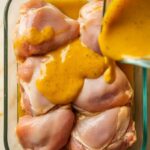 marinade being poured over raw chicken thighs in a shallow glass dish, coating them with golden sauce.