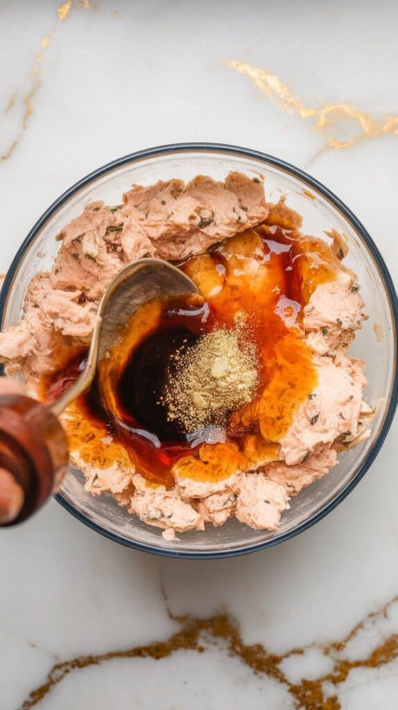 sriracha, soy sauce, and garlic powder being folded into tuna cottage cheese mix, turning slightly orange and glossy with visible seasoning.