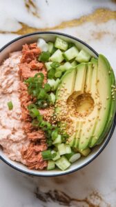 finished spicy tuna cottage cheese bowl with creamy mixture on one side, diced cucumber and fanned avocado slices on the other, green onions and sesame seeds sprinkled over the top.