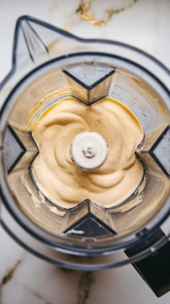 mooth lemon cottage cheese sauce mid-blend, thick texture swirling inside blender with a glossy, pale yellow finish. White marble counters with hints of gold.