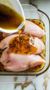 marinade being poured over raw chicken breasts in a shallow glass dish, with herbs and oil coating the surface