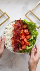 BLT cottage cheese bowl being assembled, cottage cheese, crispy bacon, halved cherry tomatoes, and chopped lettuce in neat sections.