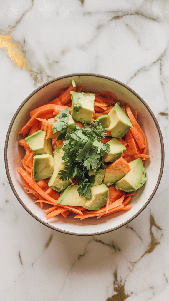finished carrot avocado salad in a serving bowl, topped with a sprinkle of chopped cilantro, avocado still intact and creamy, carrots crisp and glossy, on white marble counters