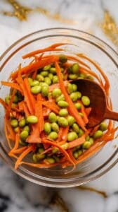 shredded carrots and edamame being tossed with soy sesame dressing in a glass bowl