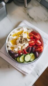 olive oil and lemon juice being drizzled over a bowl of cottage cheese, tomatoes, cucumbers, olives, and feta, finished with a dusting of dried oregano.