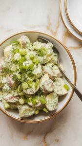 a serving bowl filled with green goddess tahini potato salad, garnished with scallions and fresh herbs, spoon resting on the side.
