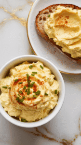 finished deviled egg salad in a bowl, garnished with smoked paprika and chopped chives. A slice of toasted bread next to it with egg salad spread on top.