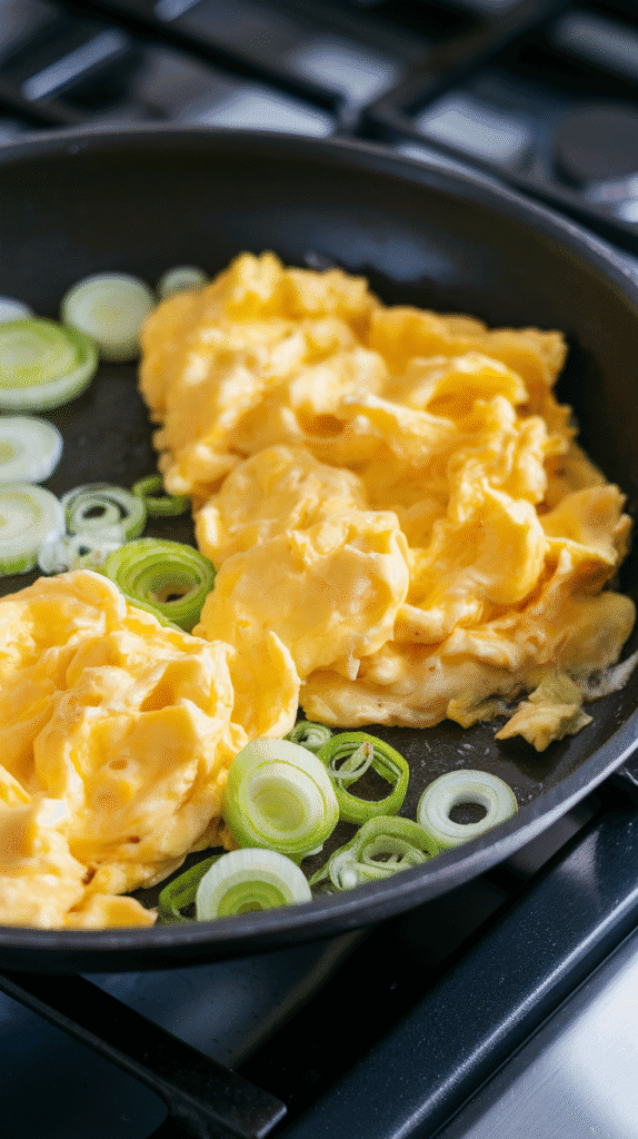 crambled eggs just starting to set on one side of the skillet, scallion whites sizzling beside them, soft folds forming.