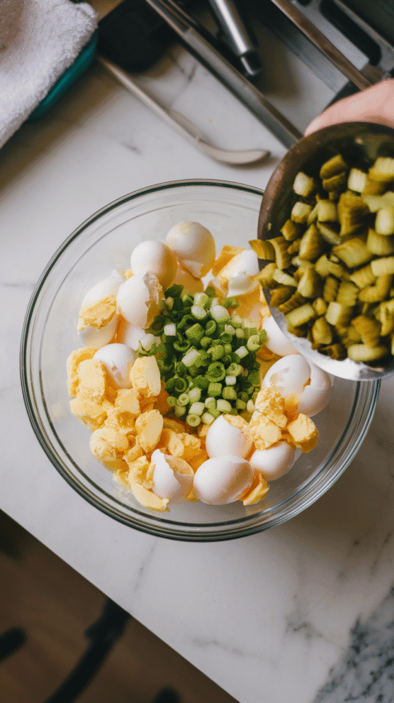 chopped pickles and green onions being added to the bowl of chopped eggs, bright green and pale yellow colors popping.