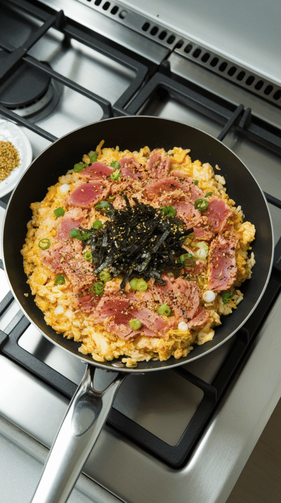 furikake seasoning and green onion being folded into hot fried rice, showing seaweed and sesame flakes over golden grains.