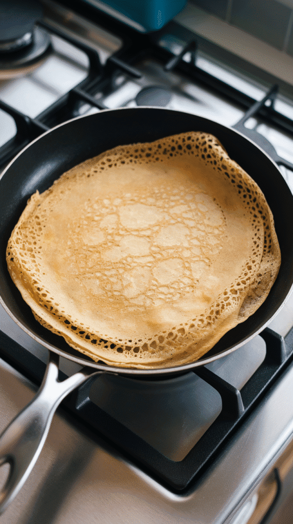 a crêpe cooking in a nonstick skillet, edges just turning golden, thin and lacy texture visible